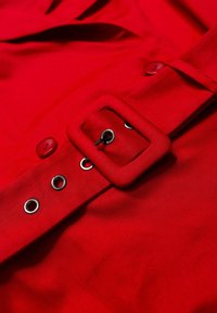 Czerwona tkanina o teksturowanej powierzchni, z szerokim paskiem z kwadratową klamrą, detalami w postaci guzików oraz srebrnymi oczkami wzdłuż boku.