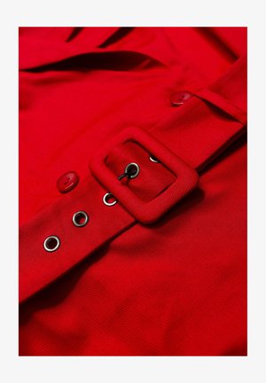 Czerwona tkanina o teksturowanej powierzchni, z szerokim paskiem z kwadratową klamrą, detalami w postaci guzików oraz srebrnymi oczkami wzdłuż boku.