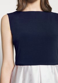Námořnická modrá bezrukávová blůza s vysokým výstřihem, spárovaná s lehkou skládací sukní. Hladká textilie a čisté linie.