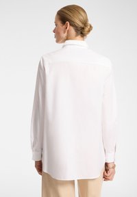 Camicia bianca a maniche lunghe con colletto, texture liscia e dettaglio con spacco sul retro, caratterizzata da polsini sartoriali e una vestibilità rilassata.
