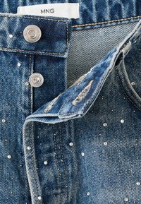 Tessuto in denim blu con embellimenti argentati. Presenta una chiusura con bottone standard e un'etichetta con la scritta "MNG" all'interno.