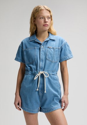 Kvinde iført lys blå kortærmet denim-onesie med forlommer og hvidt snørebælte, stående foran en ensfarvet baggrund.