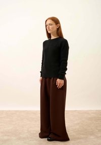 Czarny sweter z teksturowanego materiału, z długimi rękawami, w zestawieniu z szerokimi brązowymi spodniami i czarnymi butami, stylizowany na jasnym tle.