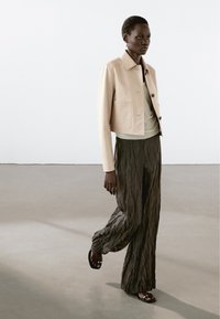 Kvinde går indendørs iført en beige cropped jakke, en lys top, mørke teksturerede brede bukser og sorte sandaler med tå-ringe.