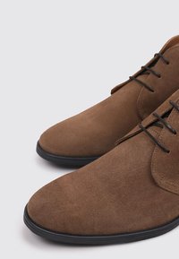 Chaussures en suède brun au design épuré, avec un bout pointu, une semelle en caoutchouc noir et un laçage noir. Texture douce et style minimaliste.