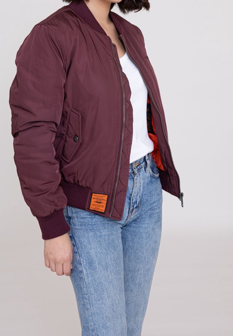 Giacca bomber borgogna con design trapuntato, polsini a coste e due tasche laterali; presenta un'etichetta arancione sul lato sinistro.