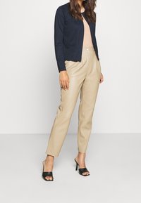 Námořnický cardigan, žebrovaný béžový top, béžové kalhoty z umělé kůže a černé sandály na podpatku. Hladké textury a přiléhavý střih.