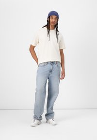 Light beige t-shirt, relaxed fit, paired with light blue straight-leg jeans. White sneakers with red and blue accents, and blue beanie.