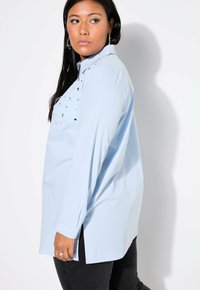Camicia a maniche lunghe azzurro chiaro con colletto, caratterizzata da dettagli decorativi sul petto e spacchi laterali. I pantaloni neri completano l'outfit.