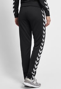 Svarta joggingbyxor med en slät textur och ett chevronmönster i vitt längs sidan. Matchas med grå fritidsskor.