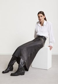 Jupe en cuir noir avec une taille haute, assortie à un chemisier blanc. Présente des accents en chaîne argentée et des bottines noires à boucles.
