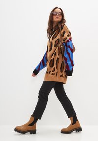 Suéter de estampado de leopardo en marrón con mangas azules y burdeos, combinado con jeans skinny negros y botines tan con paneles texturizados.