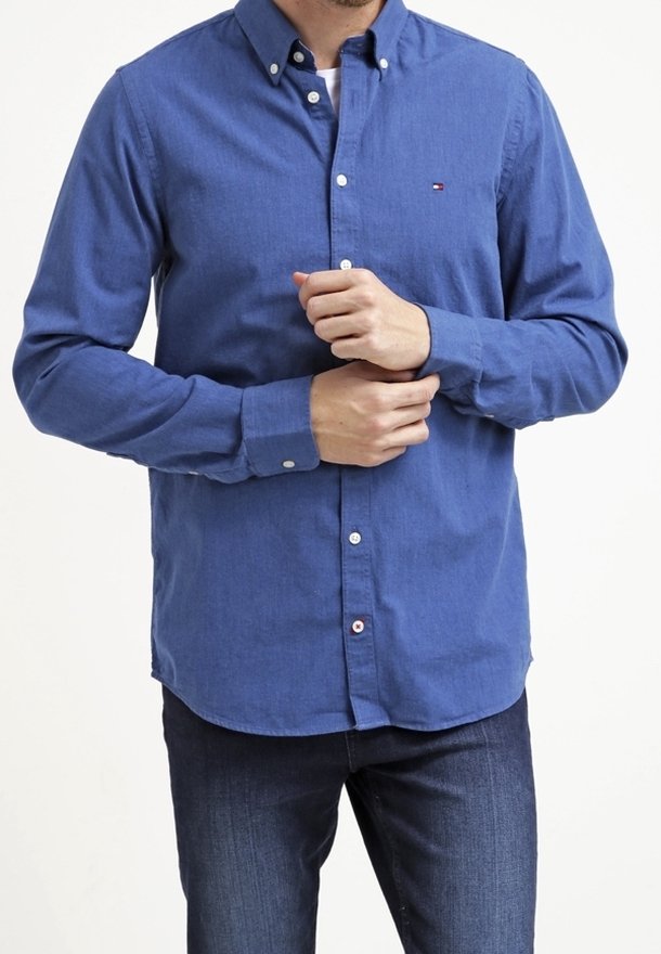 Camicia blu con colletto, maniche lunghe e orlo leggermente curvato. Presenta un piccolo logo sul petto e bottoni bianchi.