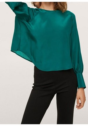 Mujer con una blusa de satén de manga larga color esmeralda y pantalones ajustados negros, levantando el brazo izquierdo mientras la blusa cae ligeramente.