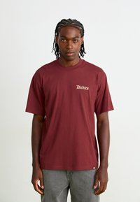 T-shirt court en coton de couleur bordeaux avec un col rond classique et un logo brodé "Dickies" en doré clair sur la poitrine gauche.
