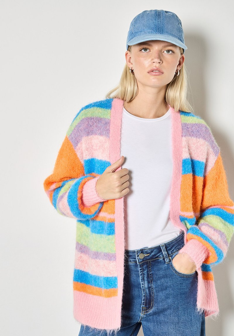 Cardigan rayé multicolore en matière duveteuse et douce, avec des nuances d'orange, de rose, de bleu et de vert. Porté sur une chemise blanche et un jean en denim.