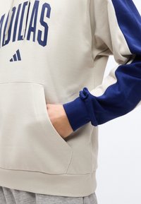 Μπεζ και ναυτικό μπλε φούτερ Adidas με τσέπη καγκουρό, χέρι μέσα στην τσέπη και γκρι φόρμες ορατές στο κάτω μέρος.