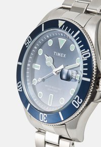 Orologio in acciaio inossidabile con lunetta blu e quadrante blu chiaro. Le caratteristiche includono indicatori luminescenti, una finestra per la data e resistenza all'acqua fino a 100 metri.