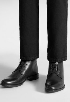 Czarne skórzane botki z gładkim wykończeniem, zaokrąglonymi czubkami i sznurowadłami, w połączeniu z ciemnymi spodniami. Widoczne subtelne przeszycia i niski obcas.