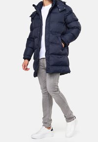 Marineblaue Steppjacke mit Reißverschluss und Druckknopfverschluss, Kapuze mit Druckknöpfen, gestepptem Textur und Seitentaschen. Getragen über einem weißen Hemd und grauen Hosen.