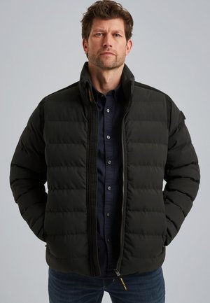 Chaqueta negra acolchada con cuello alto, cierre frontal con cremallera y bolsillos laterales. Presenta costuras horizontales y un tejido duradero. Llevada sobre una camisa azul marino.