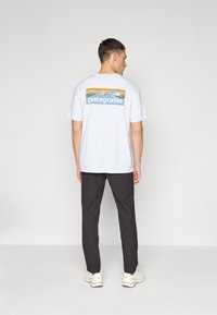 Valkoinen puuvillapaita, jossa on värikäs Patagonia-logo ja maisemasuunnittelu selässä, yhdistettynä tummiin housuihin ja vaaleisiin lenkkareihin.