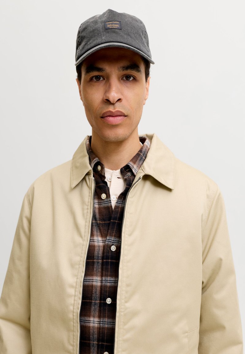 Homme portant une veste beige par-dessus une chemise à carreaux marron et une casquette de baseball grise, debout devant un fond clair uni.