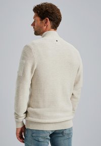 Gebreide trui in lichtbeige met geribde mouwen en zoom. Heeft een gestructureerd patroon en een patch op de linkermouw.