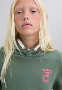 Sudadera verde con cuello ajustable mediante cordón, presenta acentos en rosa y un diseño de taza de café bordado en el pecho. Textura de tela suave.
