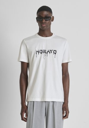 Homme portant des lunettes de soleil rectangulaires noires, un t-shirt blanc avec le texte "MORATO" en lettres coulantes, et un pantalon gris clair, debout devant un fond uni.