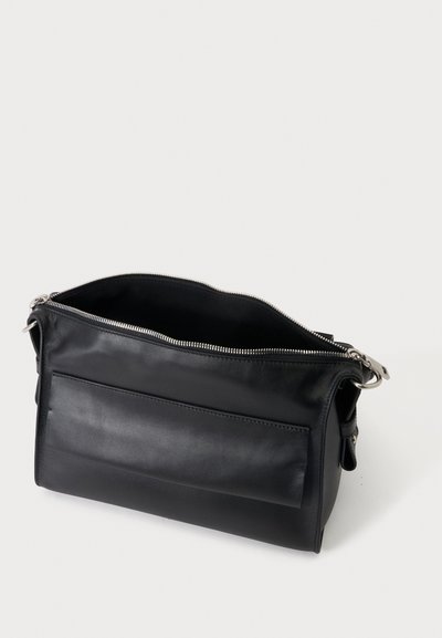 Sac bandoulière en cuir noir avec poche avant, ouverture zippée sur le dessus et ferrures métalliques argentées sur un fond clair uni.