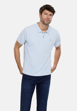 Hombre de pie, vistiendo una camiseta tipo polo de manga corta azul claro y jeans azul oscuro, con las manos apoyadas cerca de los bolsillos, fondo neutro.