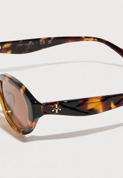 Lunettes de soleil ovales en écaille de tortue avec verres bruns et logo Tory Burch doré sur la branche contre un fond blanc.