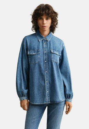 Femme aux cheveux bouclés portant une chemise en denim bleu à boutons avec des poches poitrine et un jean bleu assorti, debout devant un fond uni.