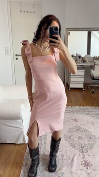 Abito rosa aderente con dettagli a fiocco sulle spalle, spacco laterale e texture liscia. La modella indossa stivali neri al ginocchio. Sfondo neutro.