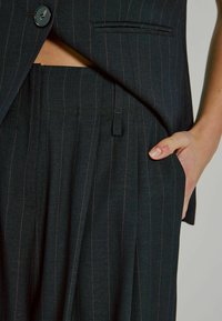 Completo nero a righe con gilet aderente e pantaloni a gamba larga. Il tessuto ha una texture liscia con una vita visibile e tasche laterali.