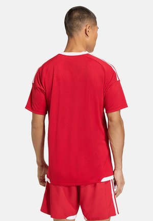 Mand iført en rød kortærmet sportstrøje og matchende shorts med hvide stribedetaljer, set bagfra mod en ensfarvet baggrund.