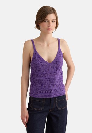 Giovane donna che indossa un top senza maniche viola lavorato a maglia con scollo a V profondo e jeans blu scuro, in piedi con una mano in tasca.