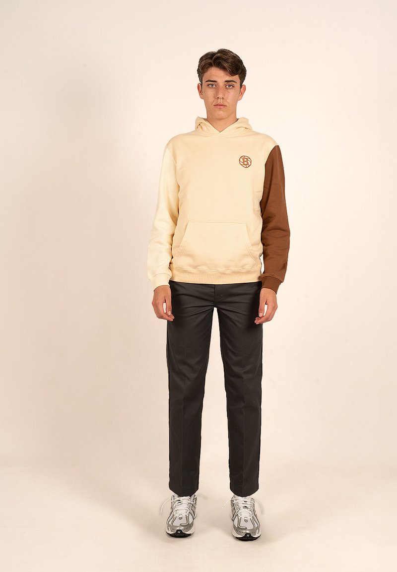 Sudadera con capucha en crema y marrón, con un bolsillo frontal y detalle de logo. Combinada con pantalones negros y zapatillas deportivas grises.