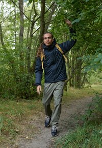 Person, der auf einem Weg im Wald geht, trägt eine schwarze isolierte Jacke, olivfarbene Cargo-Hosen und schwarze Schuhe, mit einem gelben Rucksack.