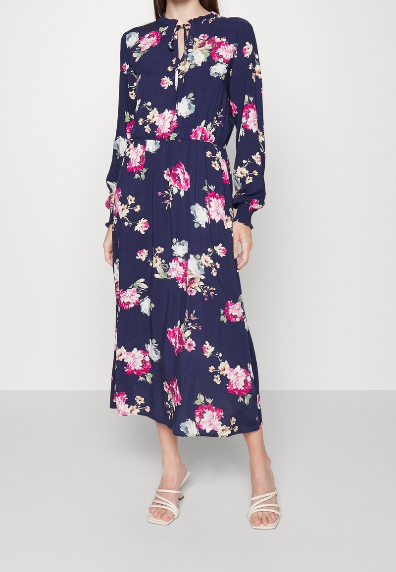 Robe bleu marine à fleurs avec manches longues, col en trou de serrure, taille cintrée et jupe fluide ornée de grands motifs floraux roses et bleus. Sandales blanches.