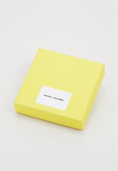 Gul firkantet boks med mat finish, der har en hvid etiket med sort tekst, der siger "MARC JACOBS." Ingen synlige mønstre eller hardware.