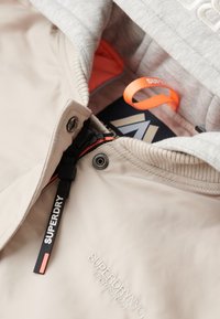 Beige jakke med grå hætte, der har lynlås lukning, en orange inderside og et sort mærke med "SUPERDRY" logo. Tekstureret stof med ribbede manchetter.