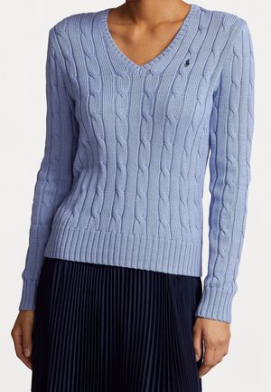 Vaaleansininen palmikkoneuleinen V-kauluksinen paita henkilöllä, joka pukeutunut tummaan pliseerattuun hameeseen, ylävartalo ja kädet näkyvissä.