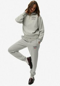 Grijze sweatshirt met ritssluiting en logo, gecombineerd met bijpassende joggers met zijstrepen en logo bij de zak. Model draagt bruine schoenen.
