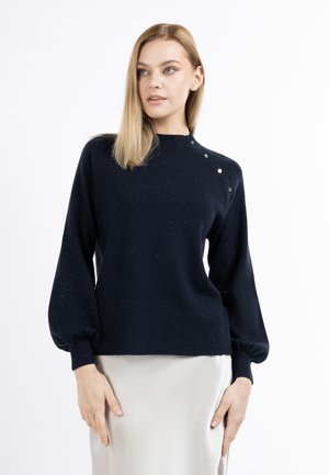 Blondynka ubrana w czarny sweter z długim rękawem z guzikami na ramieniu oraz białą spódnicę, spogląda lekko na bok.