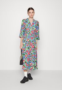 Vestido midi floral em azul, com padrões em verde, vermelho e rosa. Decote em V, mangas três quartos, barra em camadas, combinado com ténis pretos.