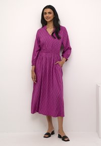 Ružičasta maxi haljina sa uzorkom, dugih rukava, v-izrezom, strukirana i s bočnim džepovima. Nosiva s crnim sandalama na blok petu.