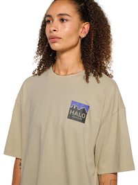 Kobieta z kręconymi włosami w beżowym, oversize T-shircie z niebiesko-czarnym logo "HALO" na piersi, patrząca lekko w lewo.