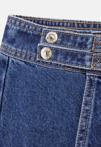 Denimbukse med en talje i blå kunststof, to sølvknapper, kontrasterende tan syninger og en let glans, som viser tekstur- og finishdetaljer.